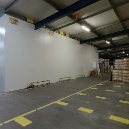 Polconstruction - Wnętrze hali magazynowej z nowo postawioną ścianą z płyt warstwowych, widoczne oznaczenia na ścianie i słupach oraz składowane towary w oddali.
