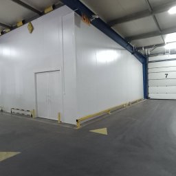 Polconstruction - Wnętrze hali magazynowej z białą ścianą z płyty warstwowej, dwuskrzydłowymi drzwiami i żółtymi odbojnicami przy podłodze, widoczna brama segmentowa z numerem 7 i palety z towarem.