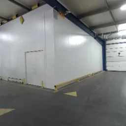 Wnętrze hali magazynowej z białą ścianą z płyty warstwowej, dwuskrzydłowymi drzwiami i żółtymi odbojnicami przy podłodze, widoczna brama segmentowa z numerem 7 i palety z towarem.