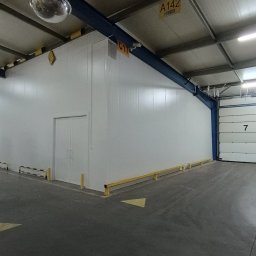 Polconstruction - Wnętrze hali produkcyjnej z zamontowaną chłodnią z płyt warstwowych, widoczne zabezpieczenia słupów i bramy, oświetlenie sufitowe i palety z towarem.