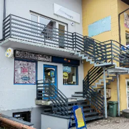 Zewnętrzne schody metalowe z poręczami prowadzące do lokalu usługowego, widoczne także balustrady na balkonie budynku.