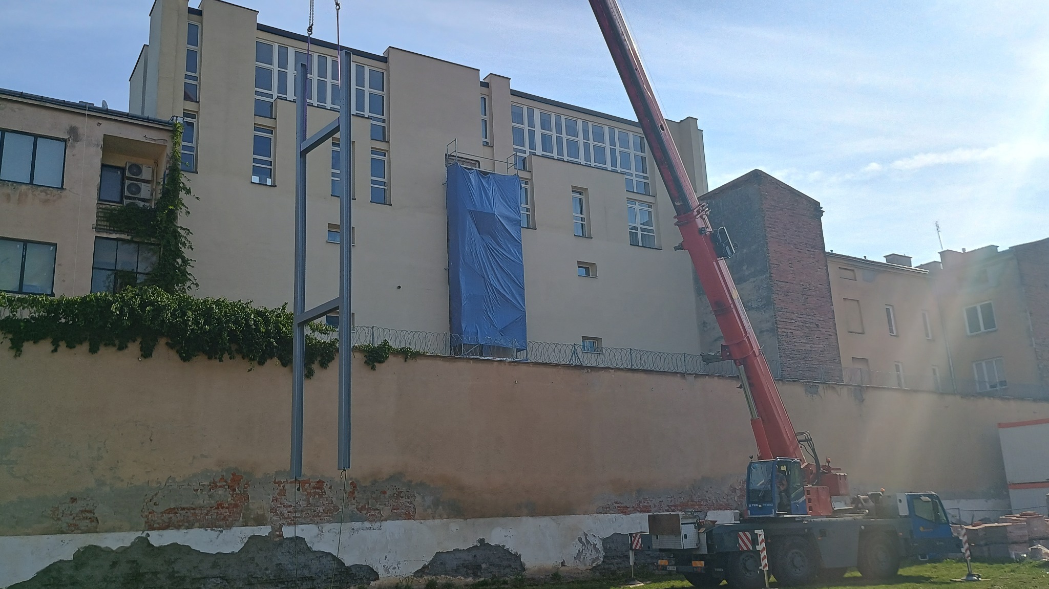 Montaż stalowej konstrukcji przy użyciu dźwigu na tle budynku z odsłoniętą cegłą i pnącą się roślinnością.