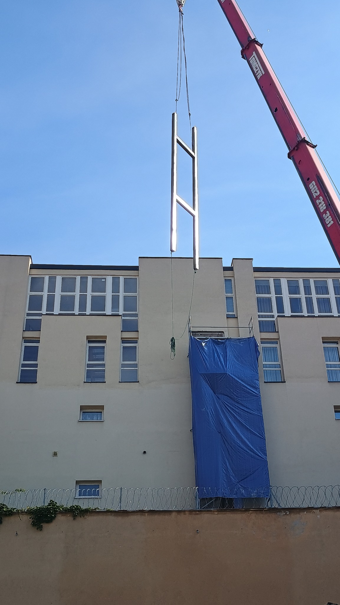 Montaż stalowej konstrukcji przypominającej literę H za pomocą dźwigu na tle budynku z niebieską plandeką ochronną.