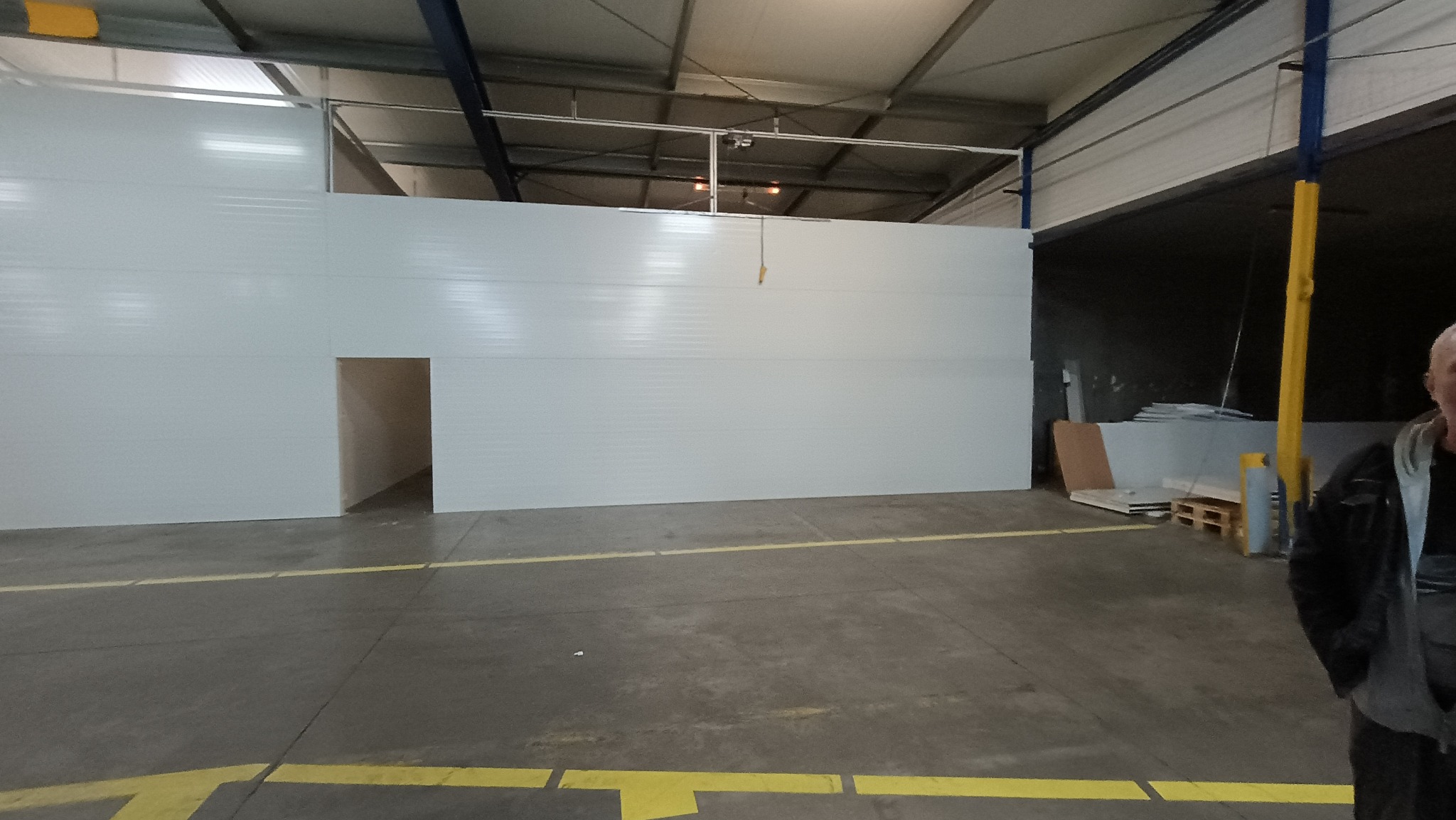 Wnętrze hali produkcyjnej z zamontowanymi białymi płytami warstwowymi i wyciętym otworem drzwiowym, widoczne żółte linie na podłodze, w tle składowane płyty i fragment sylwetki mężczyzny.