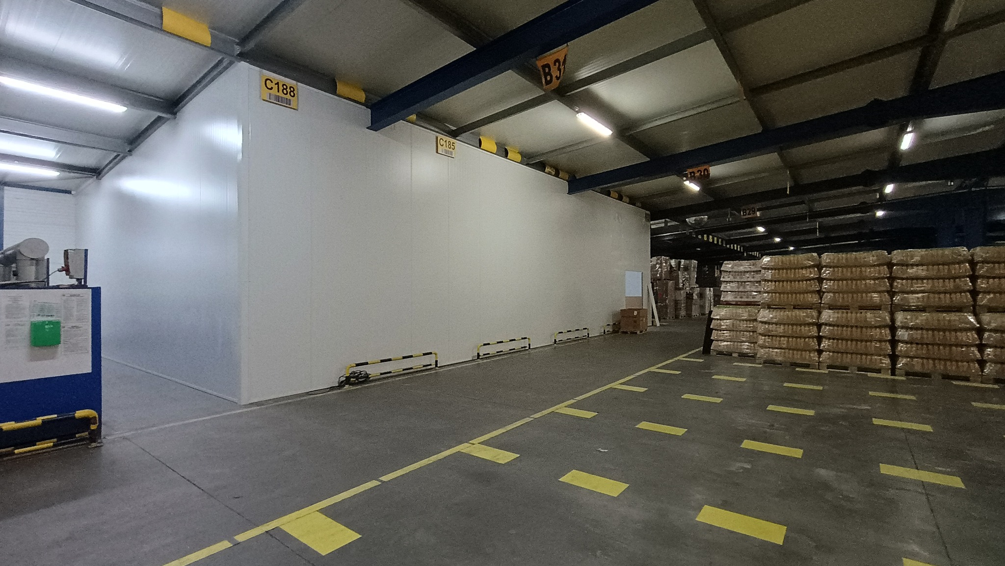 Wnętrze hali magazynowej z nowo postawioną ścianą z płyt warstwowych, widoczne oznaczenia na ścianie i słupach oraz składowane towary w oddali.