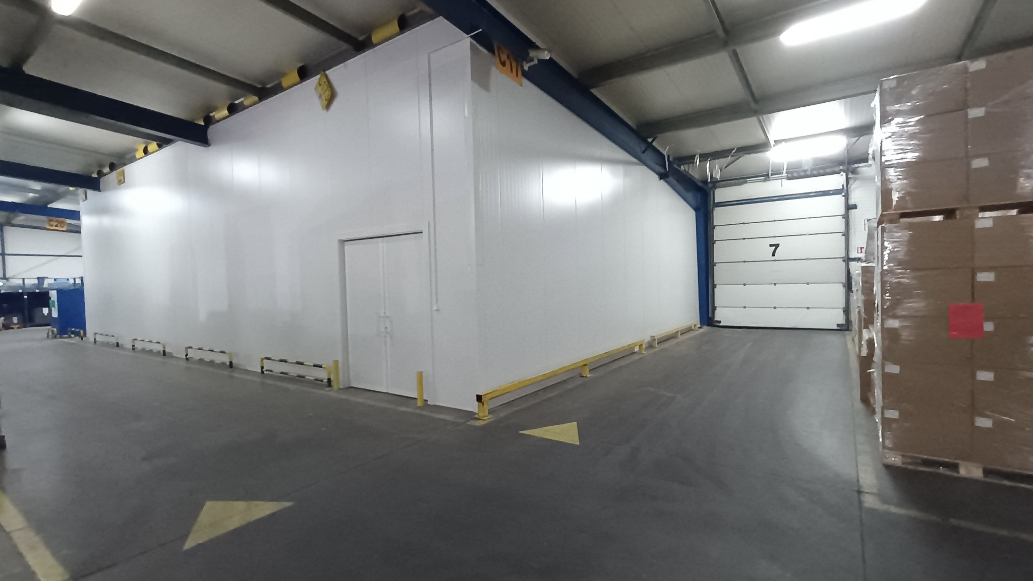 Wnętrze hali magazynowej z białą ścianą z płyty warstwowej, dwuskrzydłowymi drzwiami i żółtymi odbojnicami przy podłodze, widoczna brama segmentowa z numerem 7 i palety z towarem.