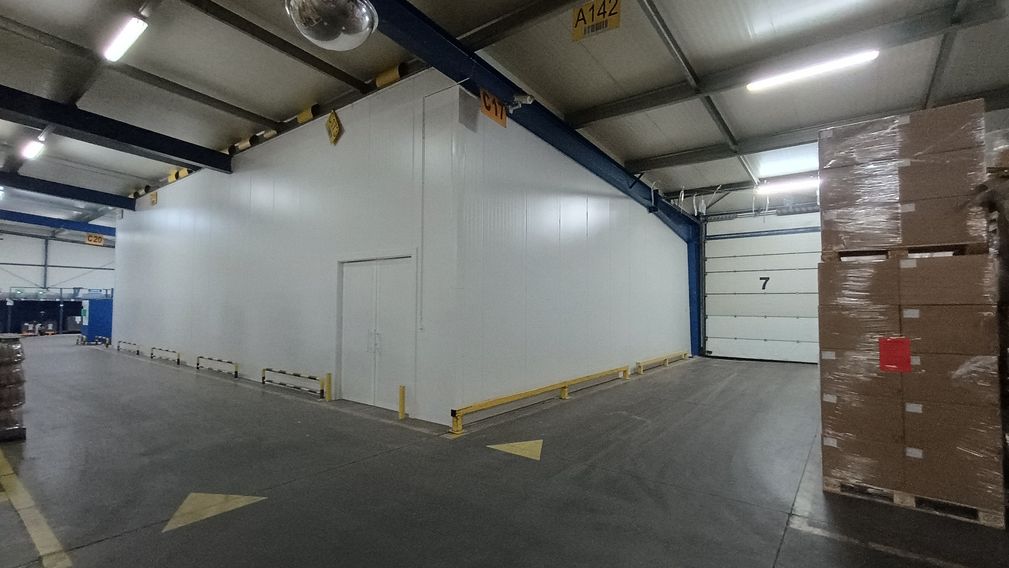 Wnętrze hali produkcyjnej z zamontowaną chłodnią z płyt warstwowych, widoczne zabezpieczenia słupów i bramy, oświetlenie sufitowe i palety z towarem.