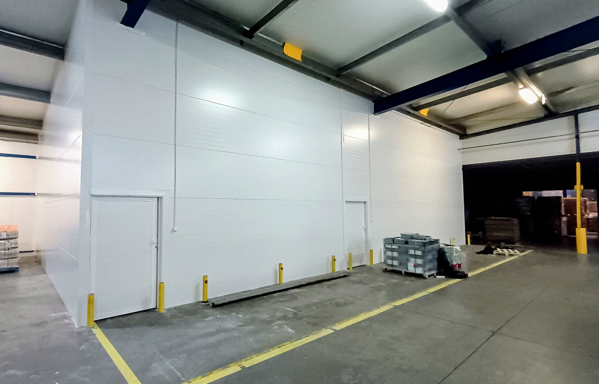 Wnętrze hali magazynowej z białymi ścianami z płyt warstwowych, dwiema parami drzwi, żółtymi słupkami ochronnymi i żółtymi liniami na podłodze.