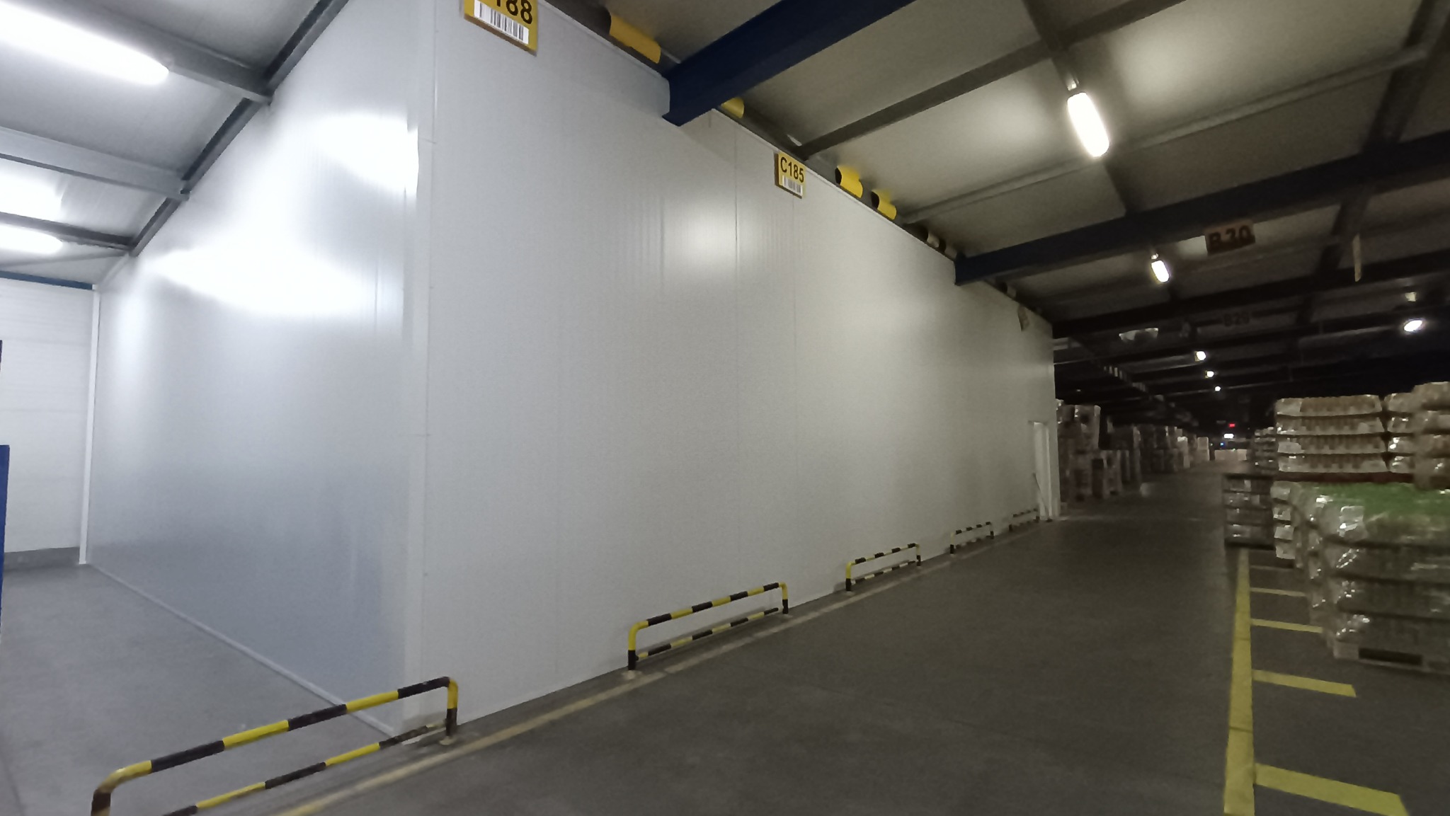 Wnętrze hali magazynowej z widocznymi ścianami z płyt warstwowych, oznaczonymi numerami identyfikacyjnymi, oraz metalową konstrukcją stropu z oświetleniem.