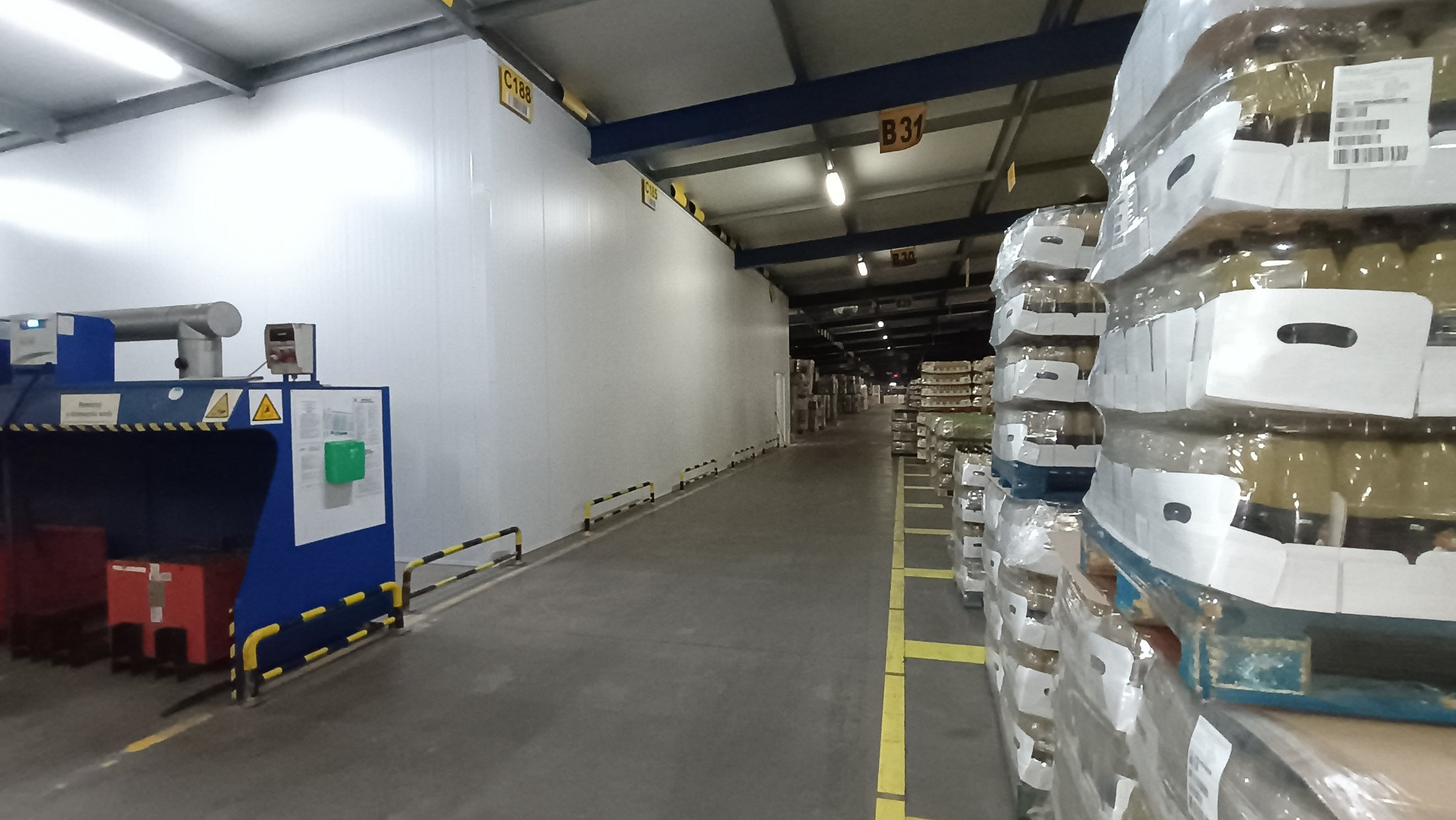 Wnętrze hali magazynowej z białymi ścianami z płyt warstwowych i rzędami palet z towarem, oświetlenie sufitowe, widoczne oznaczenia sekcji B31 i C188.