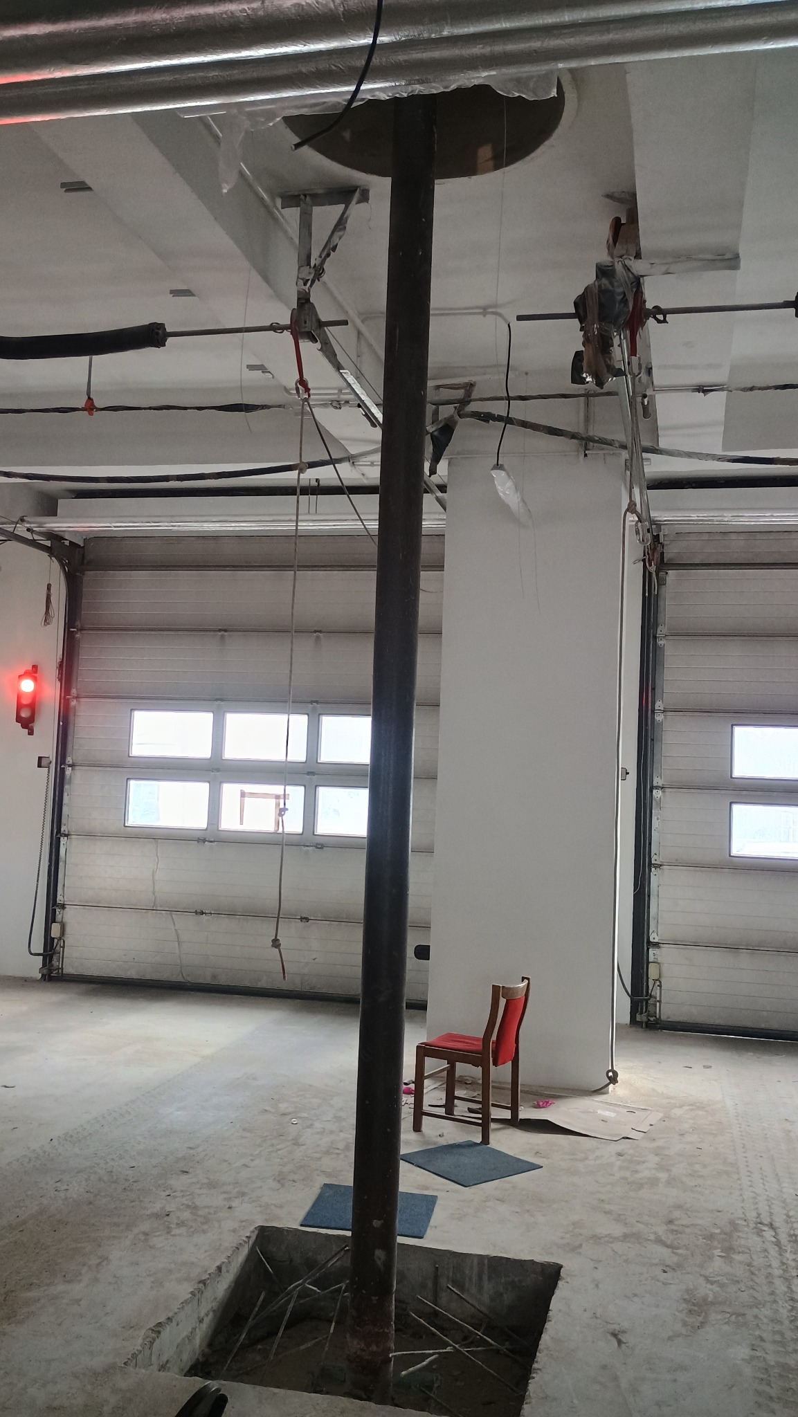 Wnętrze hali w trakcie budowy lub remontu, widoczny stalowy słup w fundamencie, instalacje pod sufitem, brama garażowa z sygnalizacją świetlną i krzesło w tle.