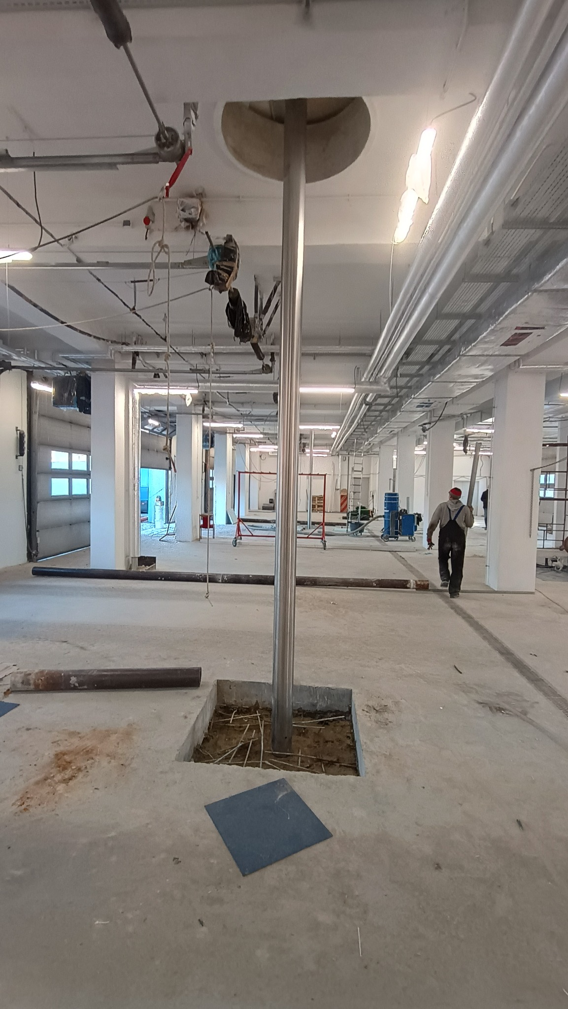 Wnętrze hali w trakcie budowy, widoczny stalowy słup w wykopie fundamentowym z wystającymi prętami zbrojeniowymi, pracownik w oddali.