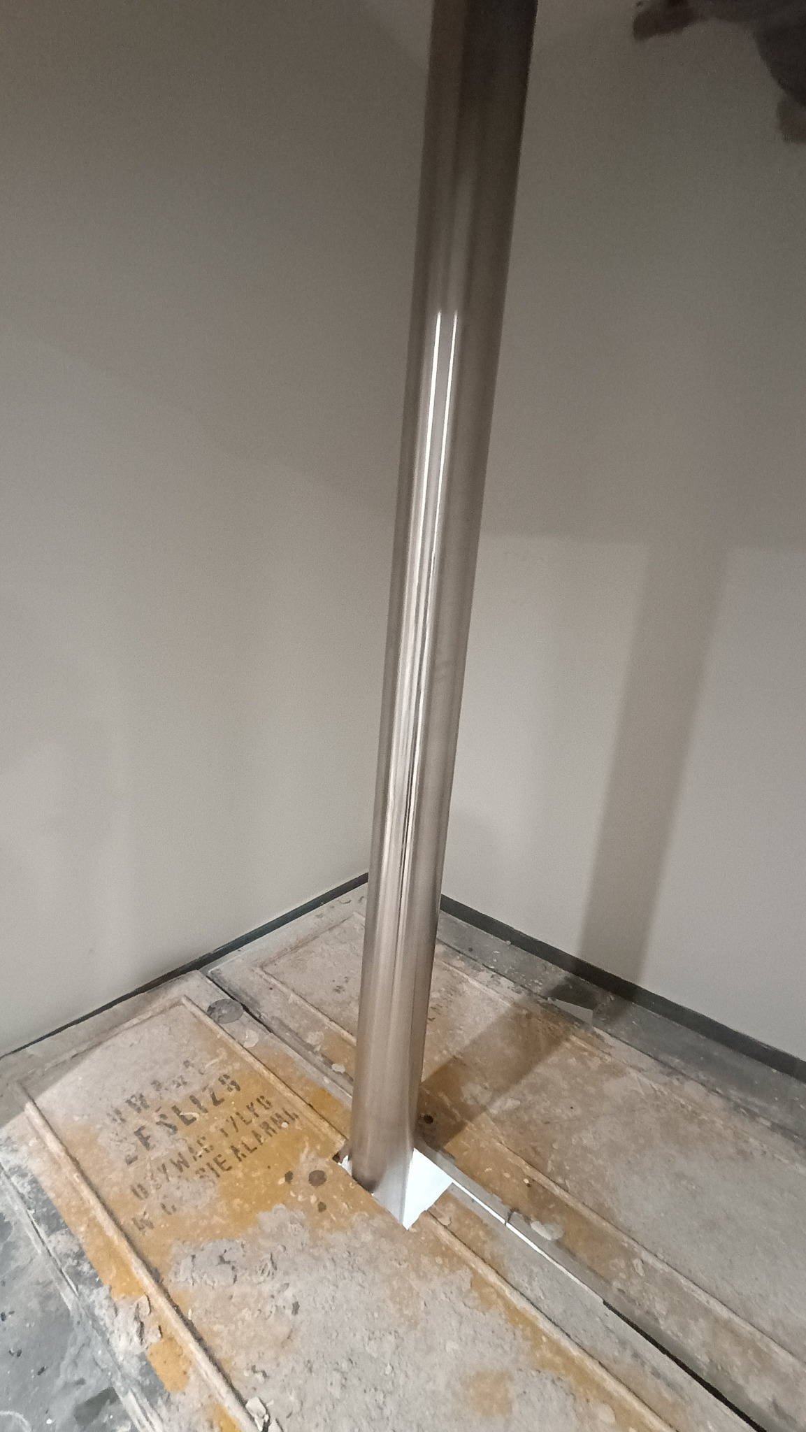 Montaż słupka poręczy ze stali nierdzewnej w podłodze z widocznymi śladami prac budowlanych i napisami ostrzegawczymi.