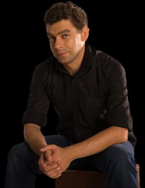 Portret mężczyzny w czarnej koszuli i dżinsach, siedzącego na brązowym stołku na czarnym tle, z rękami splecionymi przed sobą, patrzącego w obiektyw z lekkim uśmiechem.