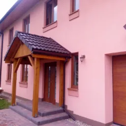 Różowy dom jednorodzinny z drewnianą konstrukcją nad wejściem, brązowym dachem i garażem. Na trawniku sterta kamieni.