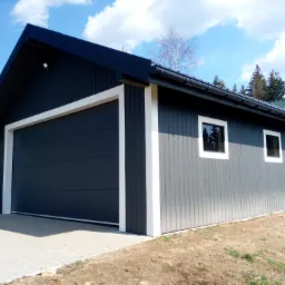 Nowoczesny garaż z szarej deski pionowej, z bramą segmentową i białymi obramowaniami, na podjeździe z kostki brukowej, w tle drzewa i błękitne niebo.