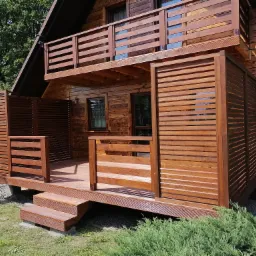 Drewniany dom z tarasem na parterze i balkonem na piętrze, otoczony zielenią; taras z drewnianymi panelami jako bariera, wejście po schodkach.