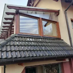 Zadaszenie tarasu z drewnianą konstrukcją i przezroczystym poliwęglanem, widok z dołu na dach pokryty dachówką ceramiczną.