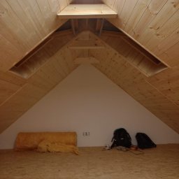 Łukasz Piotrowski Usługi Ogólnobudowlane - Poddasze z drewnianą boazerią, oknem dachowym i częściowo położoną izolacją z wełny mineralnej na podłodze z płyty OSB. Widoczne również torby i narzędzia.