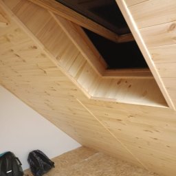 Łukasz Piotrowski Usługi Ogólnobudowlane - Drewniane wykończenie poddasza z oknem dachowym, widoczne deski boazeryjne i fragment podłogi OSB, dwa plecaki w rogu pomieszczenia.