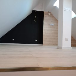 Łukasz Piotrowski Usługi Ogólnobudowlane - Wykończone poddasze w Zaskoczynie z czarną ścianą kontrastującą z drewnopodobną okładziną, jasna podłoga i białe elementy architektoniczne, widoczne okno dachowe.