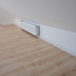 Łukasz Piotrowski Usługi Ogólnobudowlane - Poddasze po remoncie: jasne panele podłogowe, biały grzejnik zamontowany na ścianie pod skosem, biała listwa przypodłogowa.