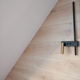 Łukasz Piotrowski Usługi Ogólnobudowlane - Ściana pod skosem wykończona panelami drewnopodobnymi, na której zamontowano nowoczesną, czarno-złotą lampę ścienną z dwoma włącznikami.