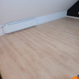 Łukasz Piotrowski Usługi Ogólnobudowlane - Poddasze z nową, jasną podłogą laminowaną imitującą drewno, białe ściany i grzejnik. Fragment czarnej ściany w rogu. Wiadro z akcesoriami do sprzątania w prawym dolnym rogu.