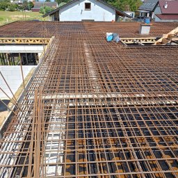 Łukasz Piotrowski Usługi Ogólnobudowlane - Zbrojenie płyty fundamentowej z widoczną siatką z prętów stalowych oraz elementami szalunku i izolacji, w tle zabudowa jednorodzinna.
