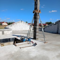 Łukasz Piotrowski Usługi Ogólnobudowlane - Wznoszony komin z bloczków betonowych, podparty stemplami budowlanymi na płycie fundamentowej. Widoczne wiaderka, szpadel i inne narzędzia budowlane. W tle wzniesione ściany z bloczków i błękitne...