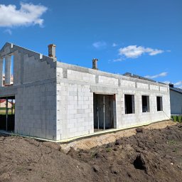 Łukasz Piotrowski Usługi Ogólnobudowlane - Surowy stan domu jednorodzinnego z bloczków betonowych, widoczne otwory okienne i drzwiowe, kominy w trakcie budowy, ocieplenie fundamentów, ziemia wokół budynku.