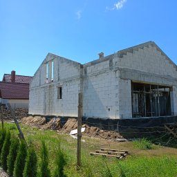 Łukasz Piotrowski Usługi Ogólnobudowlane - Surowy stan budynku z białych bloczków, widoczne otwory okienne i zbrojenia, teren wokół budynku częściowo zalany wodą, w tle dom z czerwoną dachówką.