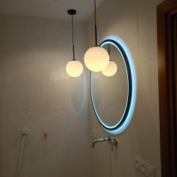 Łukasz Piotrowski Usługi Ogólnobudowlane - Nowoczesna łazienka z białymi, prostokątnymi płytkami, okrągłym lustrem z podświetleniem LED, chromowaną baterią umywalkową i trzema lampami wiszącymi z kulistymi kloszami.