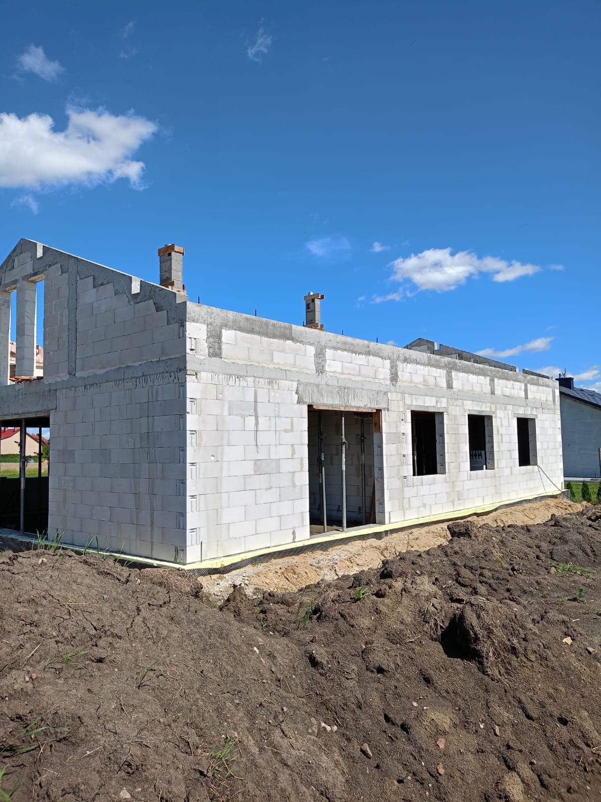 Surowy stan domu jednorodzinnego z bloczków betonowych, widoczne otwory okienne i drzwiowe, kominy w trakcie budowy, ocieplenie fundamentów, ziemia wokół budynku.