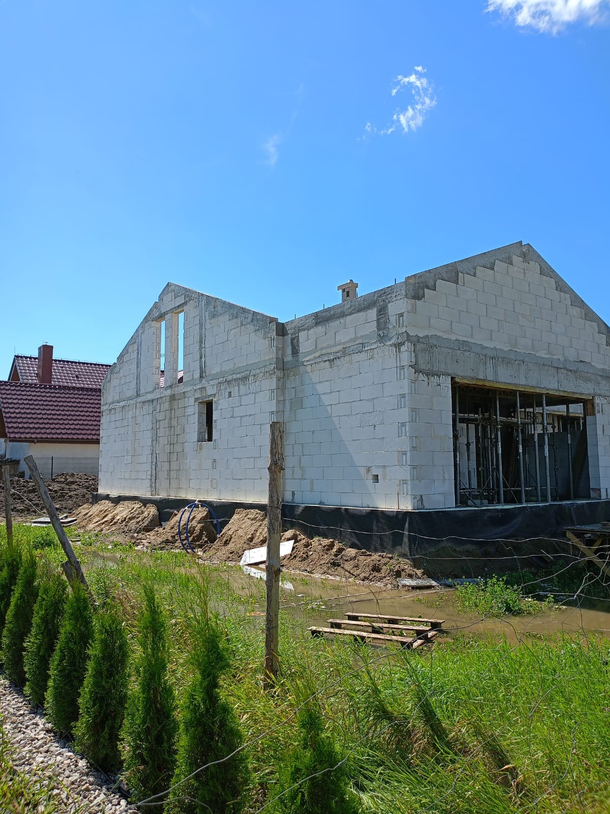 Surowy stan budynku z białych bloczków, widoczne otwory okienne i zbrojenia, teren wokół budynku częściowo zalany wodą, w tle dom z czerwoną dachówką.