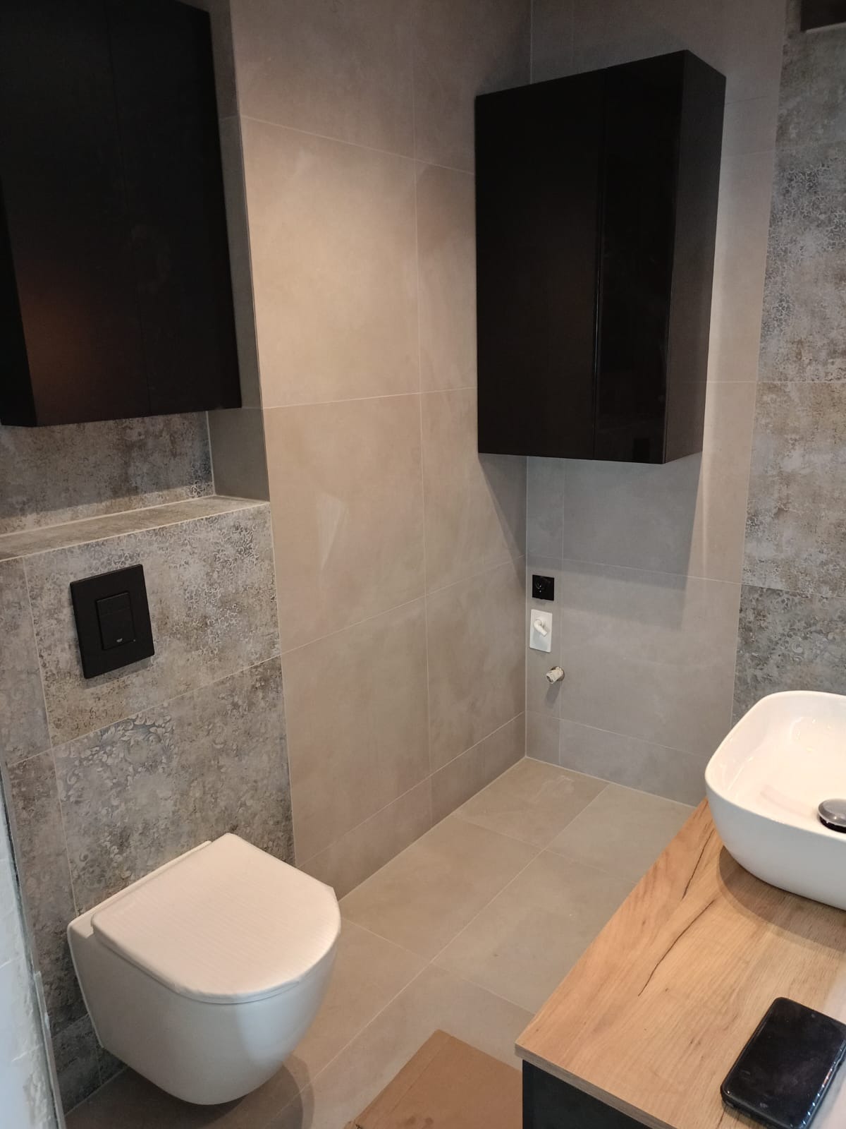 Nowoczesna łazienka z wiszącą toaletą, dwoma czarnymi szafkami, umywalką nablatową na blacie z drewnopodobnym dekorem i szarą glazurą.