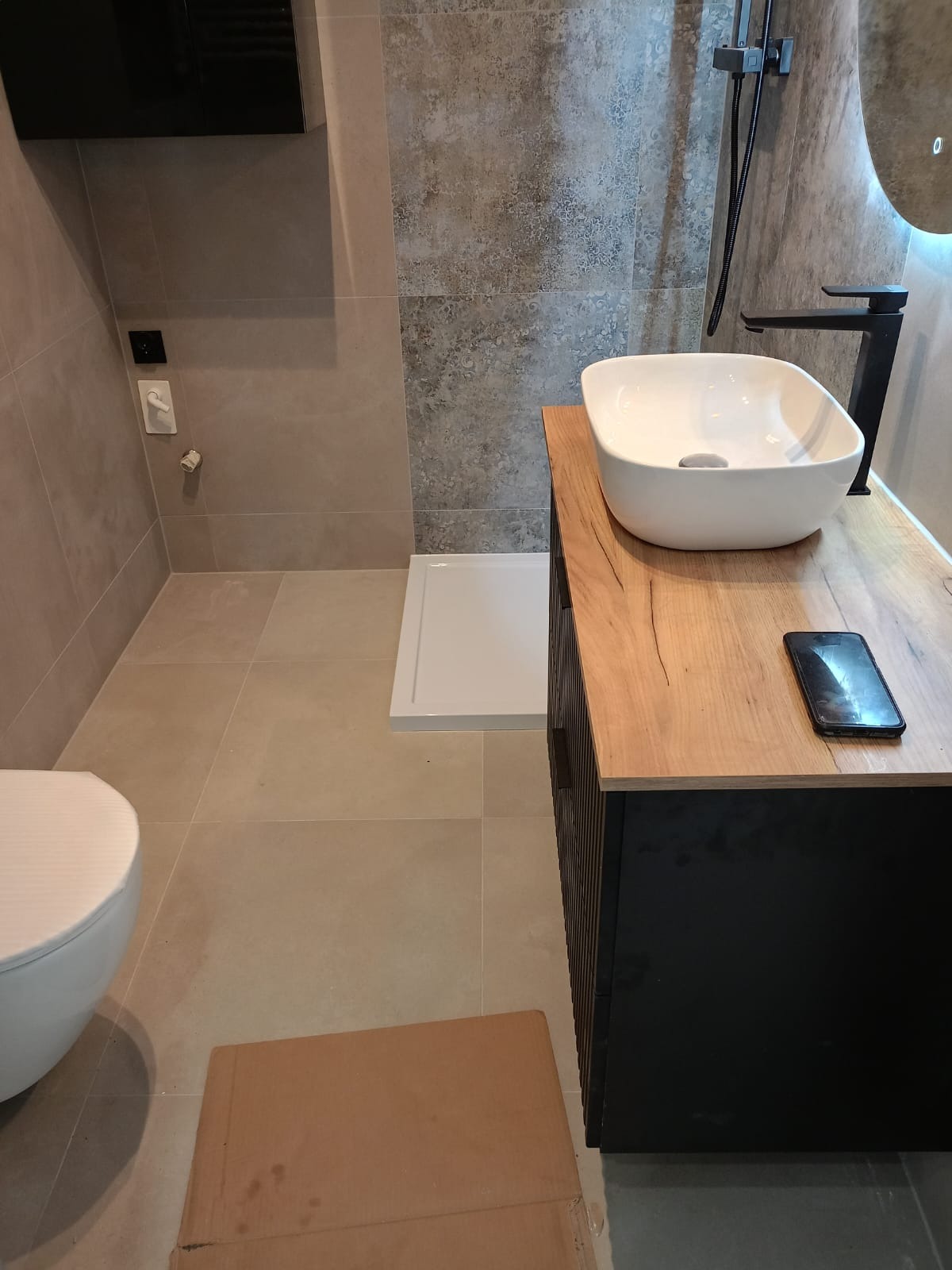 Nowoczesna łazienka z umywalką nablatową, czarną armaturą i kabiną prysznicową typu walk-in, widoczny telefon na blacie szafki umywalkowej oraz karton na podłodze.