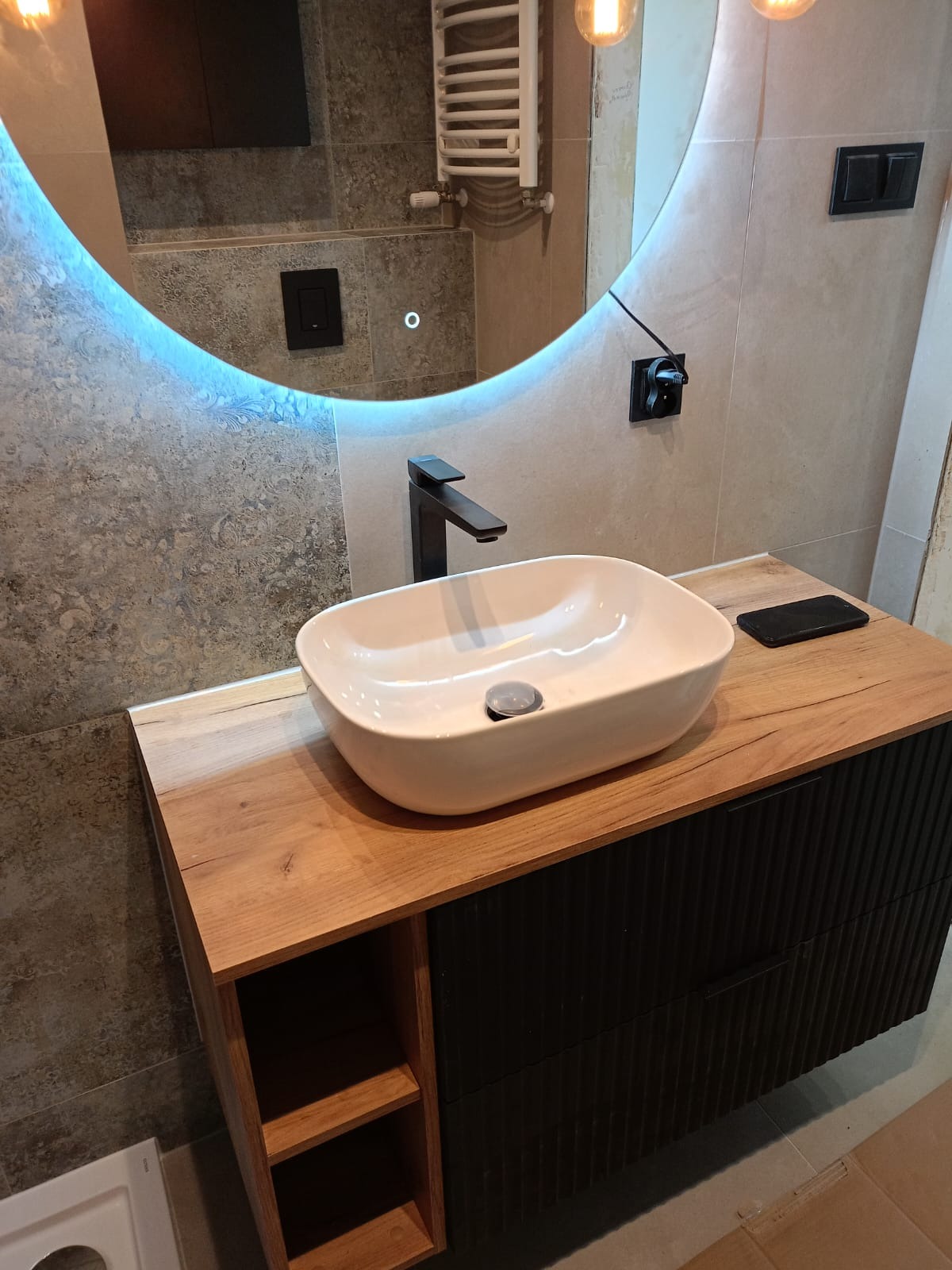 Umywalka nablatowa o owalnym kształcie, z czarną, minimalistyczną baterią, zamontowana na drewnianym blacie z szafką z ryflowanymi frontami. Nad umywalką podświetlane okrągłe lustro z widocznymi...