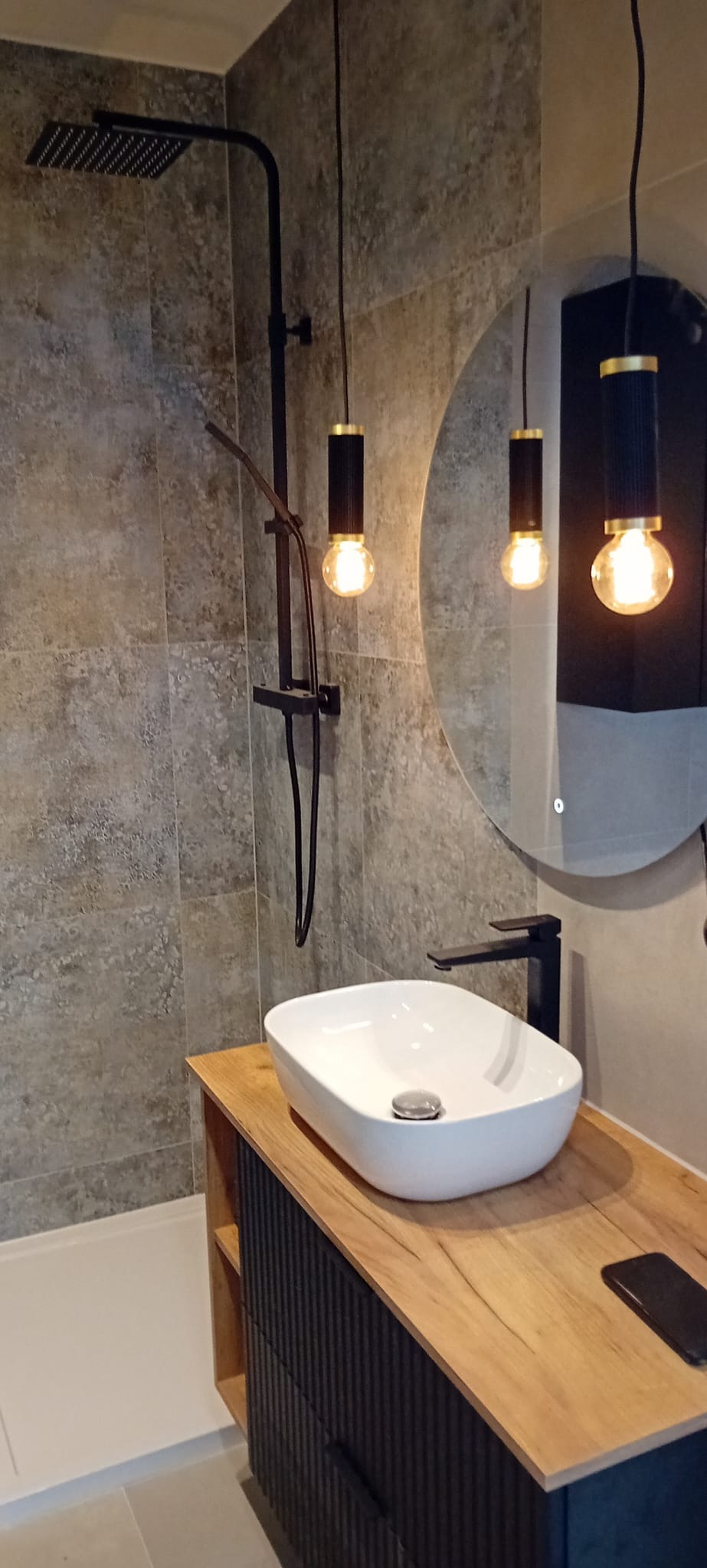 Nowoczesna łazienka z szarą płytką imitującą kamień, czarną armaturą, okrągłym lustrem z podświetleniem LED i wiszącymi lampami z żarówkami Edisona, umywalka nablatowa na drewnianej szafce...