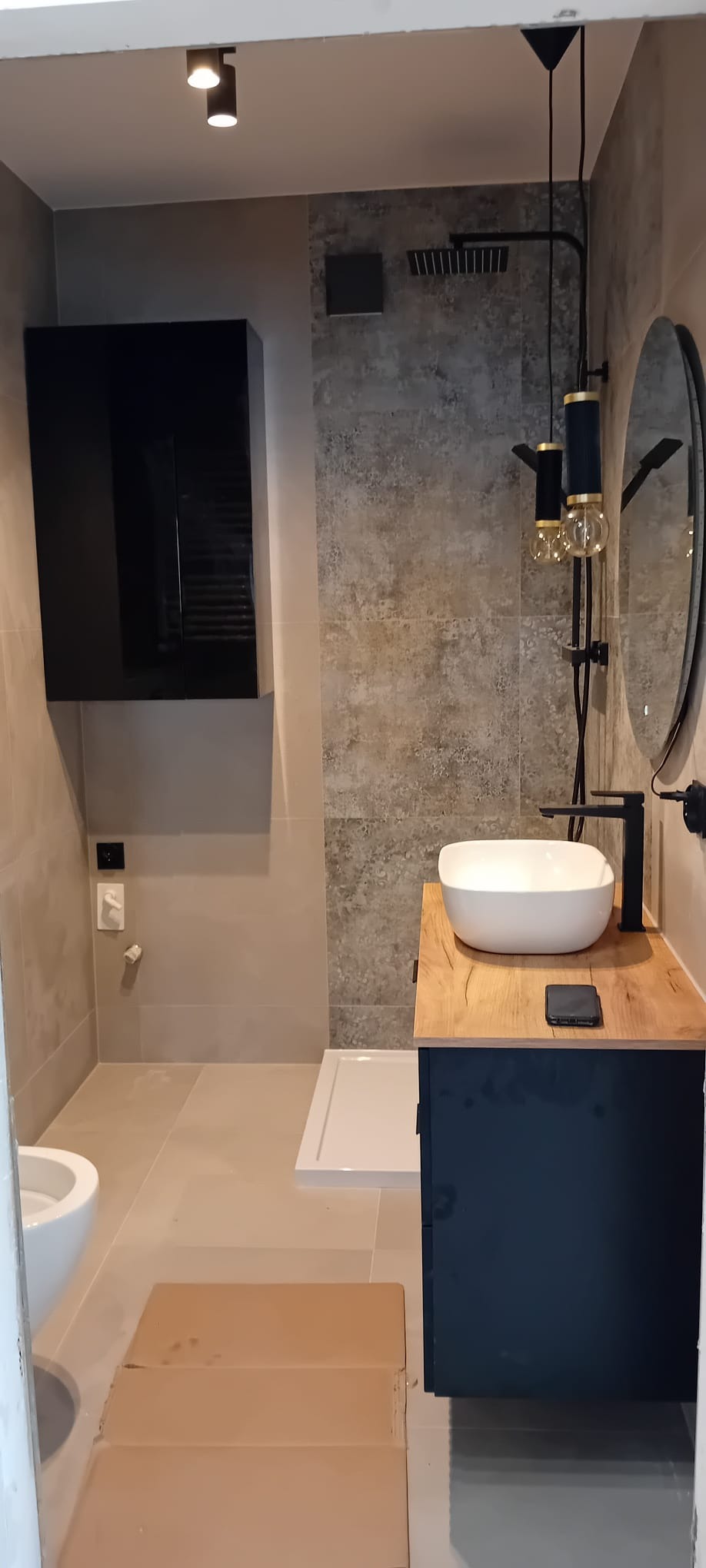 Nowoczesna łazienka z umywalką nablatową na drewnianym blacie, czarną armaturą, kabiną prysznicową typu walk-in i wiszącą szafką, oświetlona reflektorami.