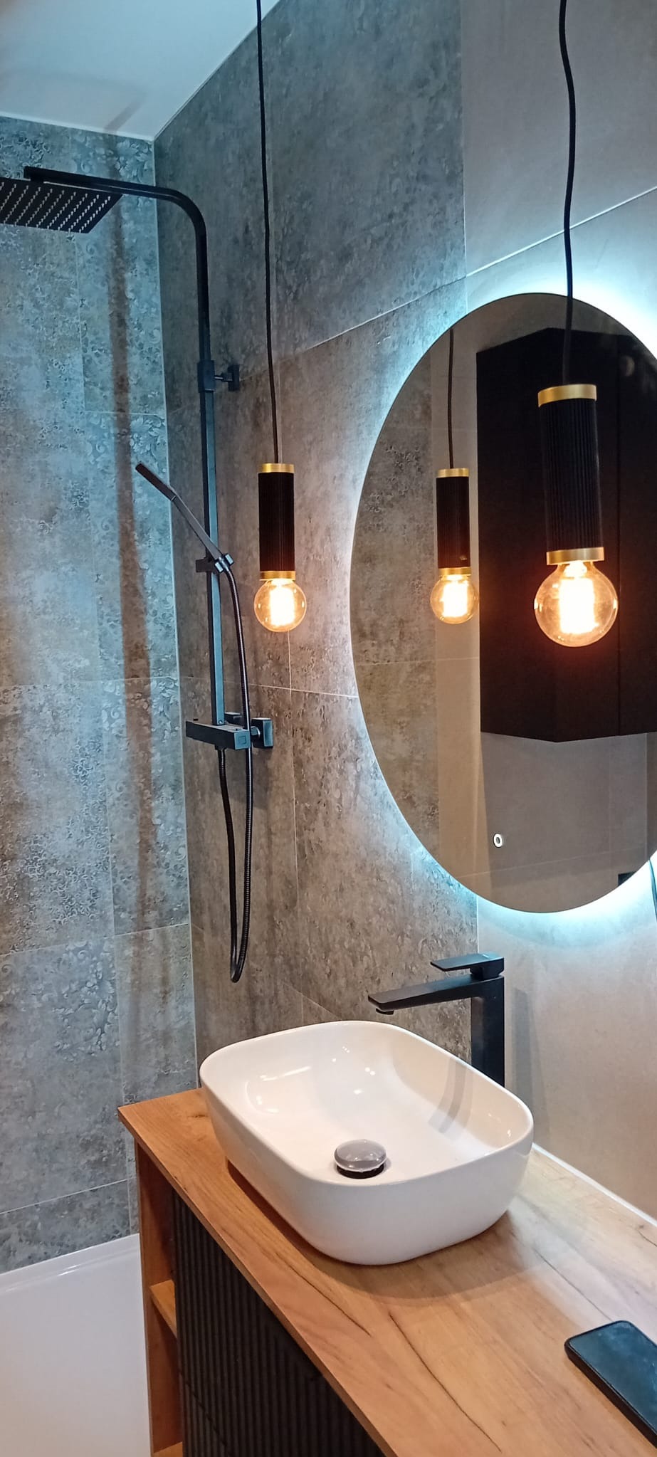 Nowoczesna łazienka z owalną białą umywalką nablatową, czarną armaturą, deszczownicą i okrągłym lustrem z podświetleniem LED oraz wiszącymi lampami z żarówkami Edisona.