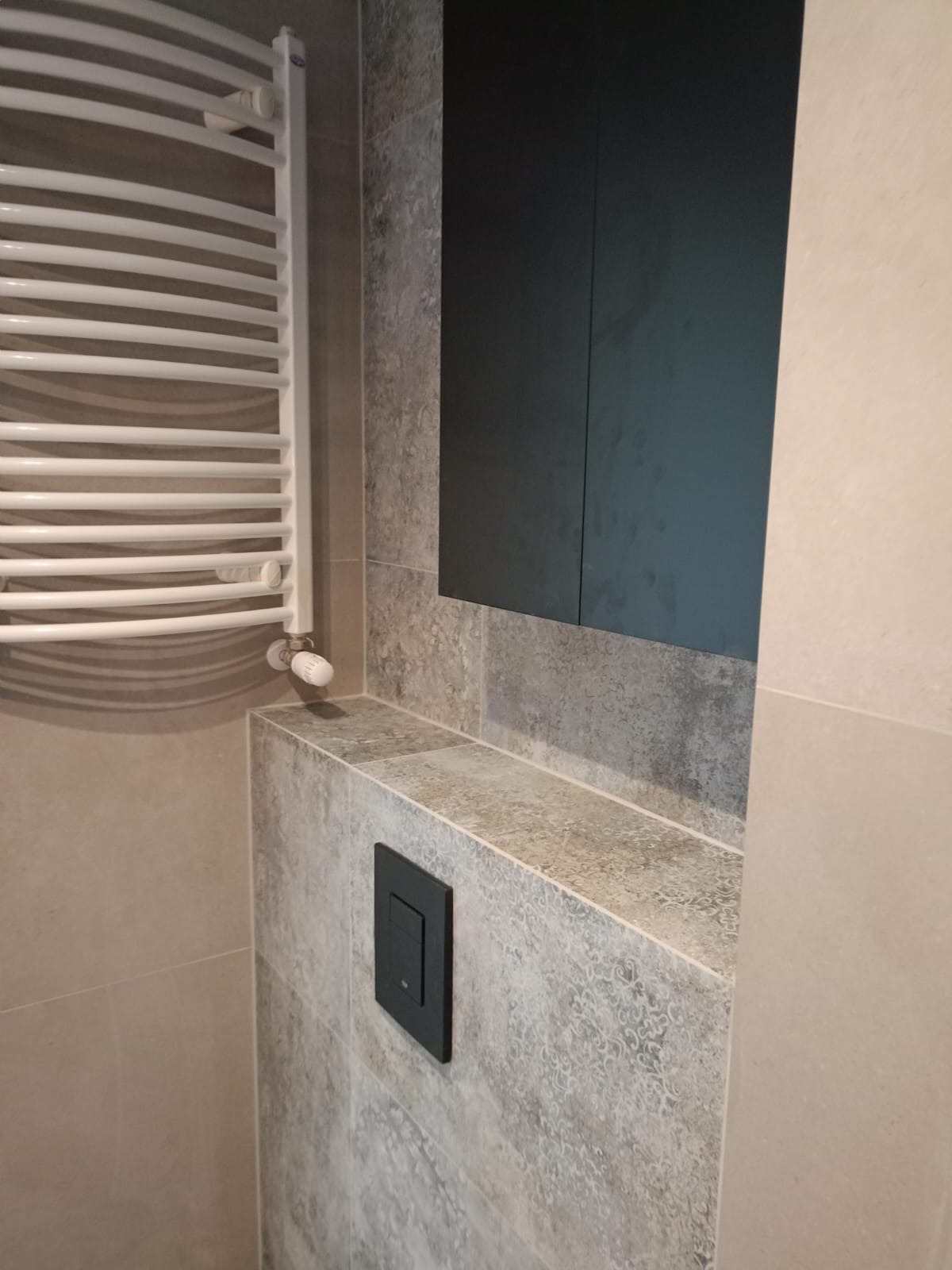 Fragment wykończonej łazienki: grzejnik drabinkowy, czarna szafka wisząca i panel sterowania spłuczką WC na tle płytek imitujących kamień.