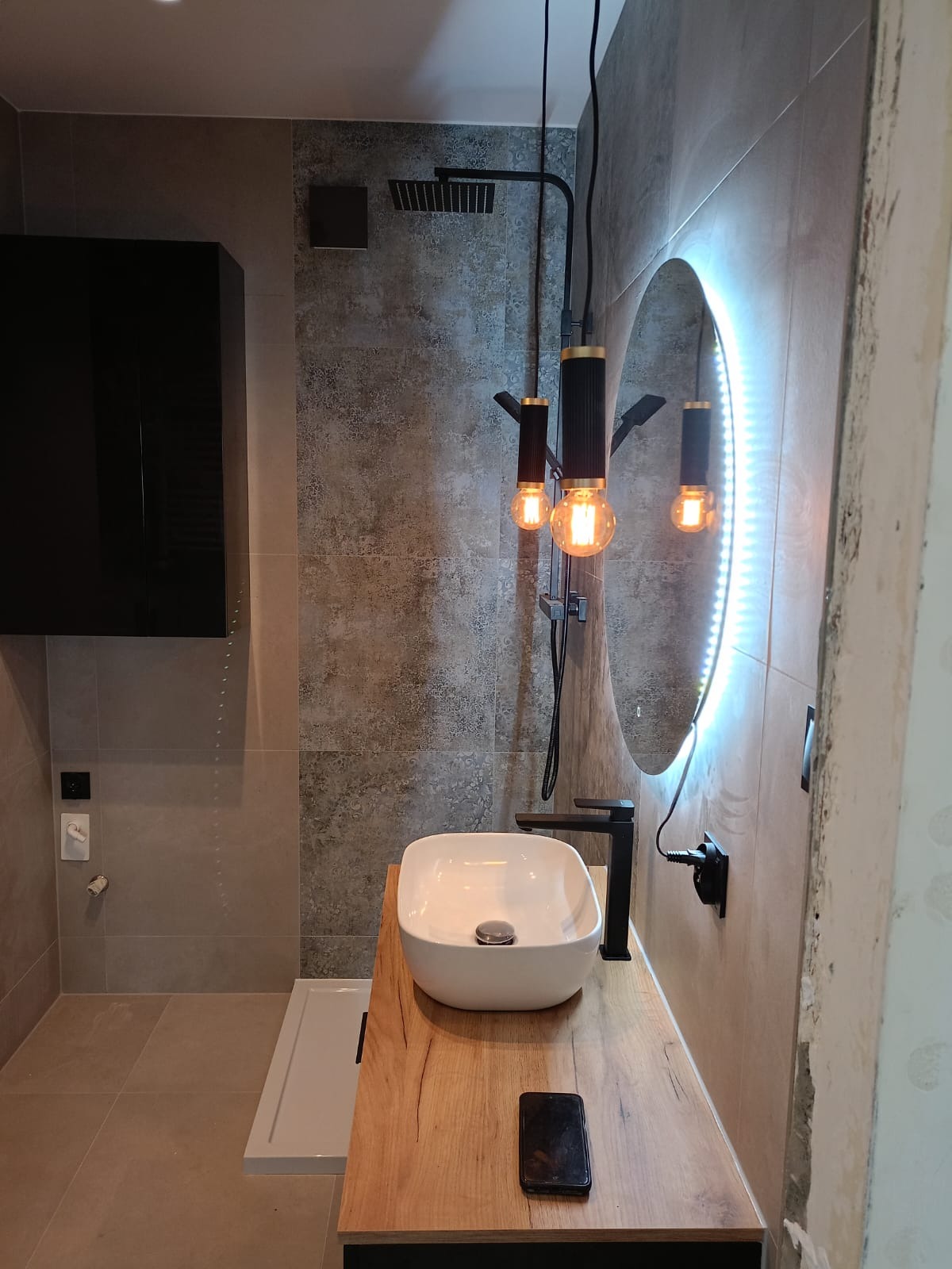 Nowoczesna łazienka z owalnym lustrem podświetlanym LED, wiszącą armaturą w kolorze czarnym, umywalką nablatową i drewnianym blatem oraz wiszącą szafką.
