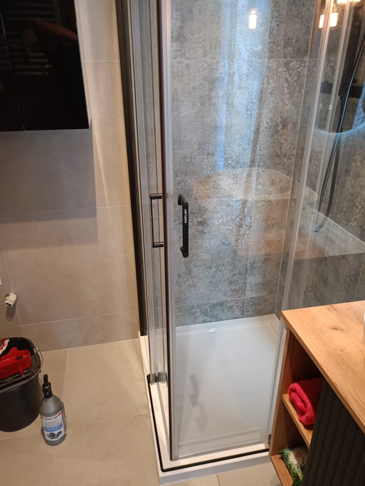 Nowoczesna kabina prysznicowa z czarnymi detalami, szklane drzwi z uchwytem firmy Sanplast, szare płytki z motywem roślinnym w tle, widoczny fragment szafki umywalkowej z drewnianym blatem...