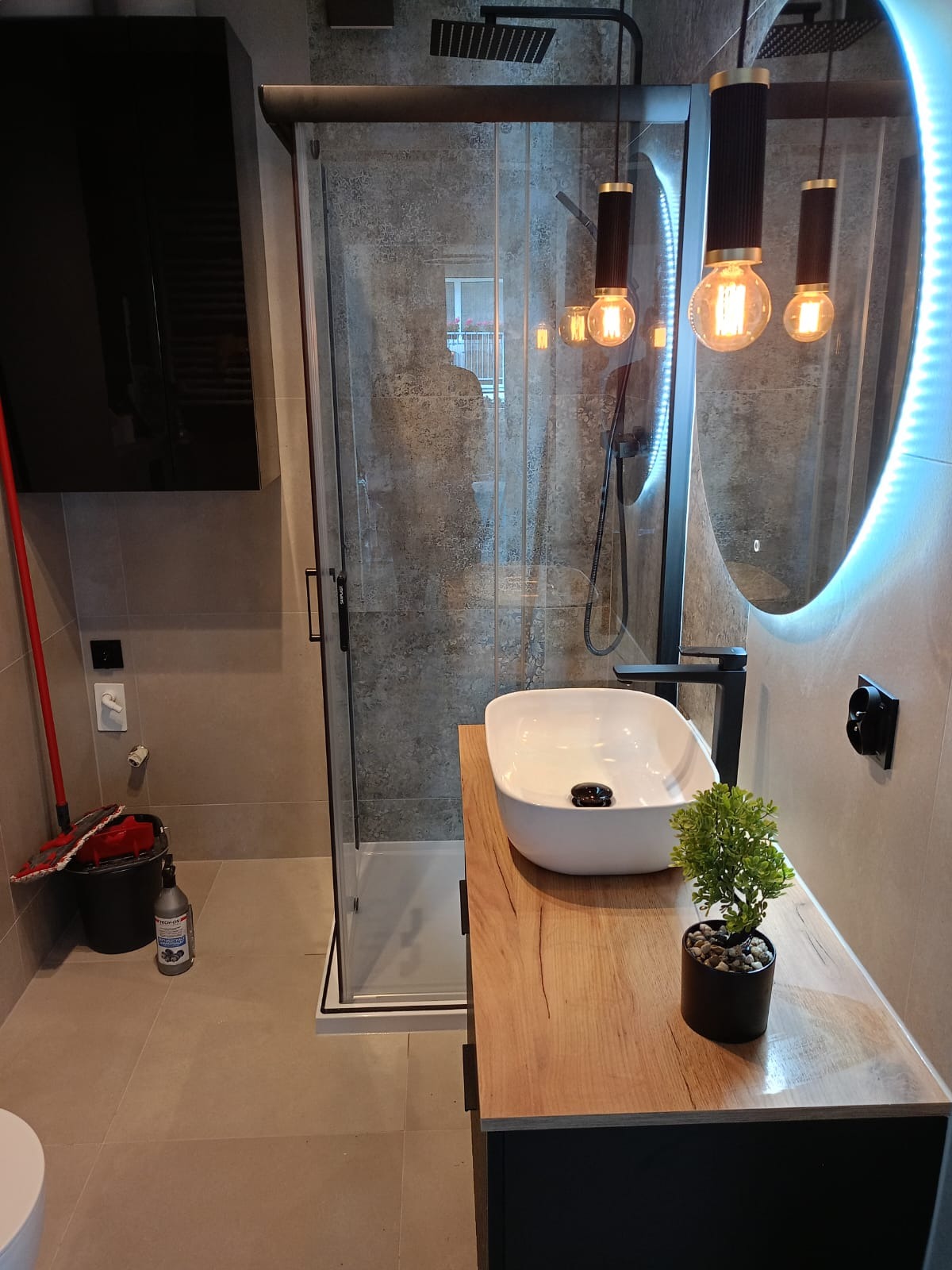 Nowoczesna łazienka z kabiną prysznicową typu walk-in, umywalką nablatową na drewnopodobnym blacie, okrągłym lustrem z podświetleniem LED i wiszącymi lampami z żarówkami Edisona