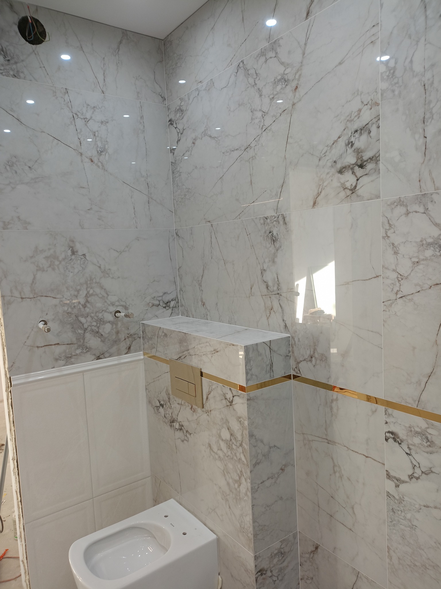 Wykończona łazienka w Zaskoczynie z marmurowymi płytkami, złotymi akcentami na obudowie stelaża WC i chromowanymi przyłączami wody, widoczne przygotowane punkty świetlne w suficie.