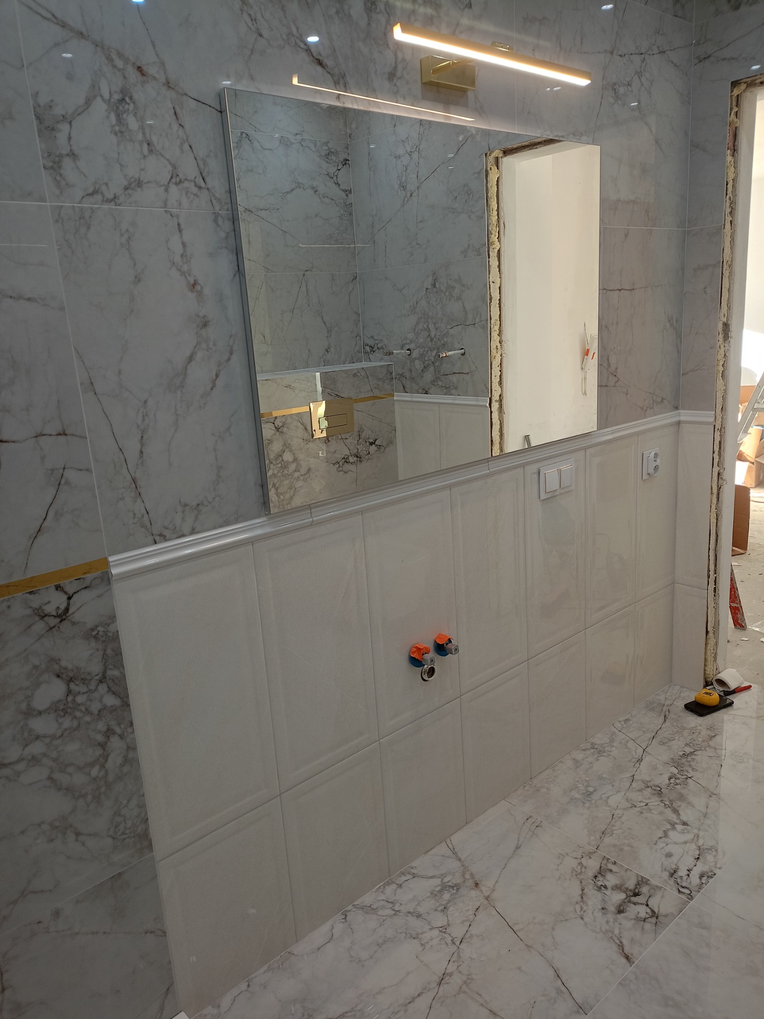 Ściana łazienki wyłożona płytkami imitującymi marmur, z zamontowanym lustrem z podświetleniem LED, fragment ściany z białymi płytkami z dekoracyjnym obramowaniem i wystającymi zaworami.