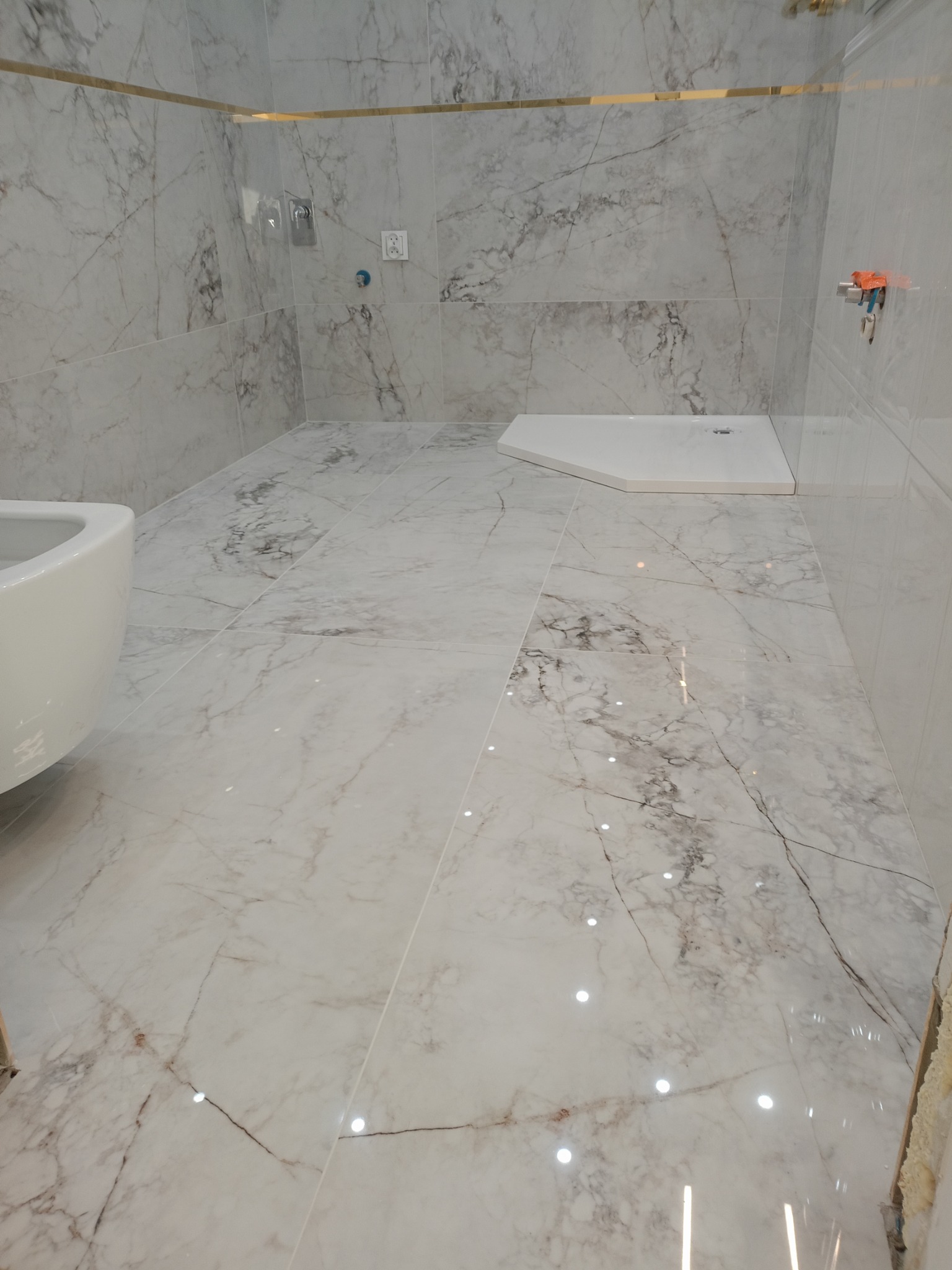 Wykończona łazienka z białymi marmurkowymi płytkami na ścianach i podłodze, z brodzikiem i częściowo widoczną toaletą.