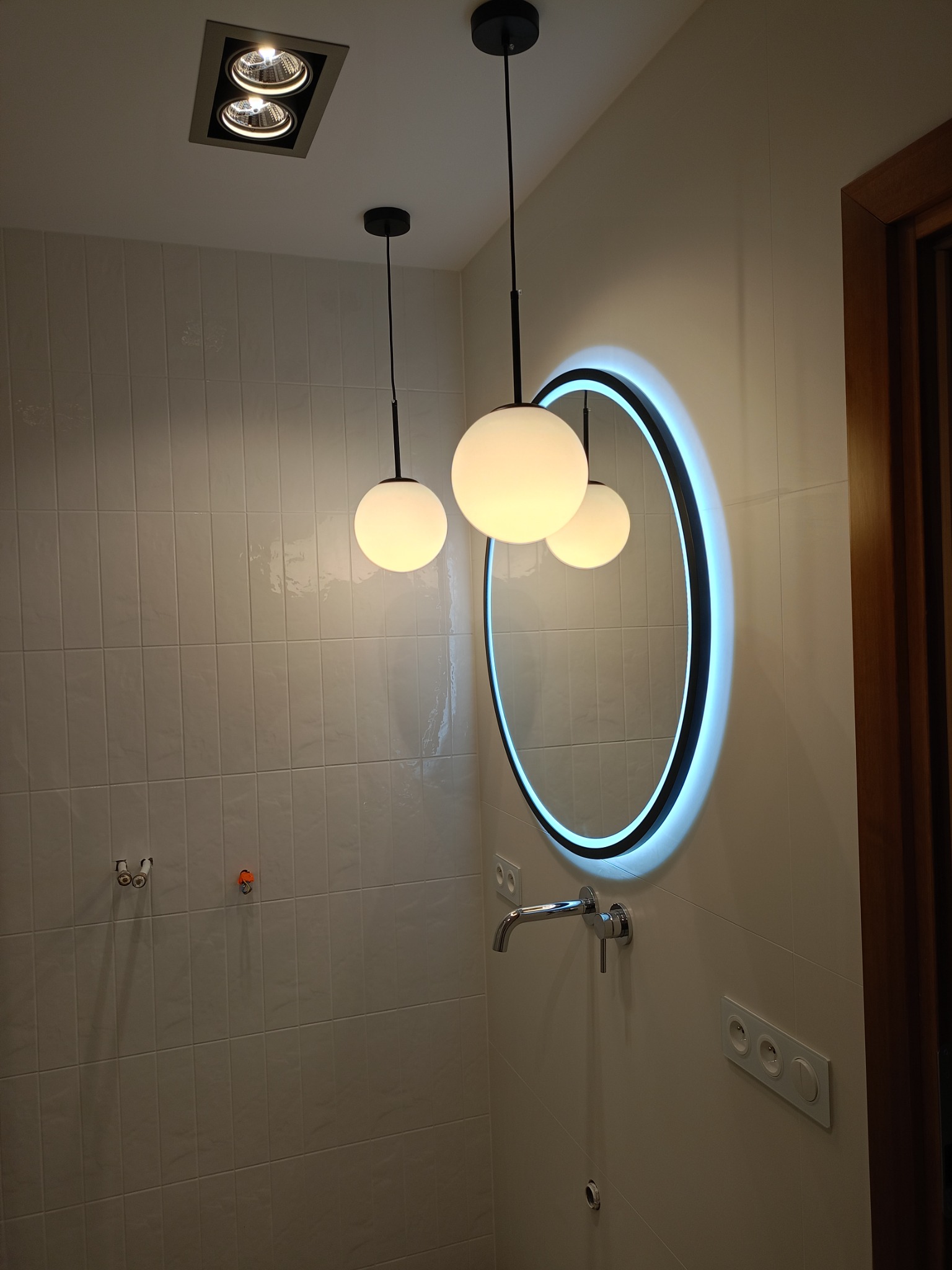 Nowoczesna łazienka z białymi, prostokątnymi płytkami, okrągłym lustrem z podświetleniem LED, chromowaną baterią umywalkową i trzema lampami wiszącymi z kulistymi kloszami.