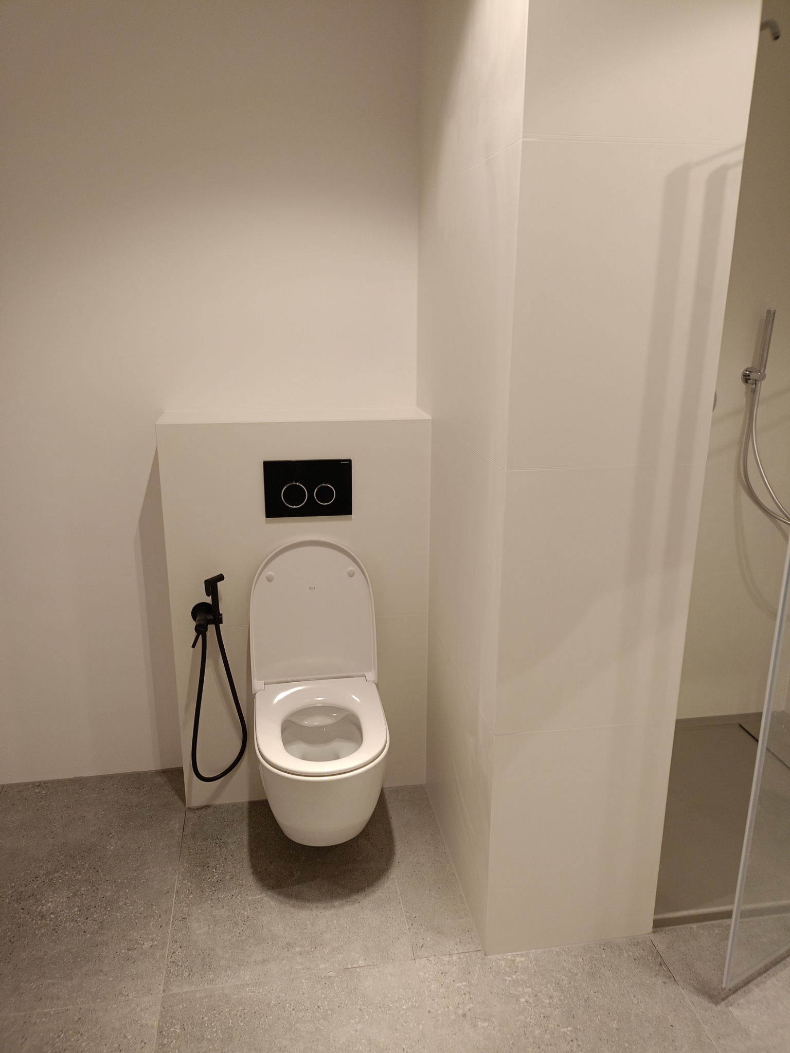 Nowoczesna łazienka z podtynkową toaletą, czarnym panelem sterującym spłukiwaniem i bidetką w kolorze czarnym, szare płytki podłogowe, kabina prysznicowa w tle.